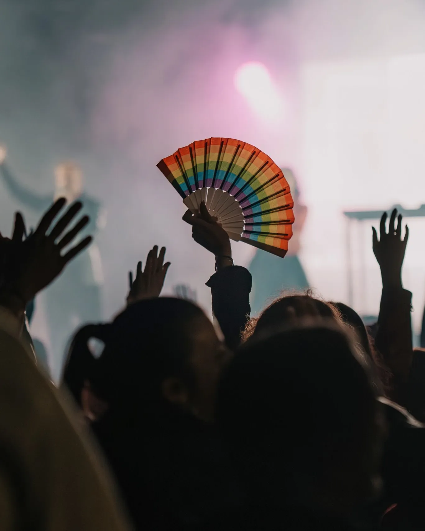 Públic assistent a un esdeveniment amb els braços aixecats mentre una persona sosté un ventall amb els colors de l’arc de Sant Martí, símbol de la diversitat LGBTIQ+.