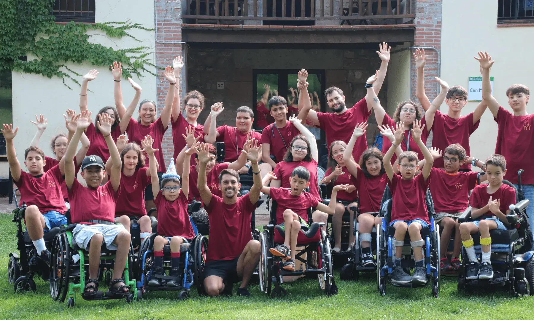 Grup d’infants i joves, alguns amb mobilitat reduïda i en cadira de rodes, juntament amb monitors i monitores, aixequen les mans en senyal d’alegria davant d’un edifici en un entorn natural.