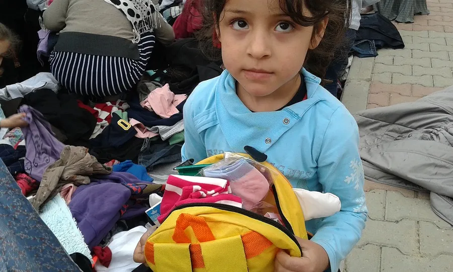 Una nena siriana mira a la càmera amb la mirada perduda, sostenint una motxilla.