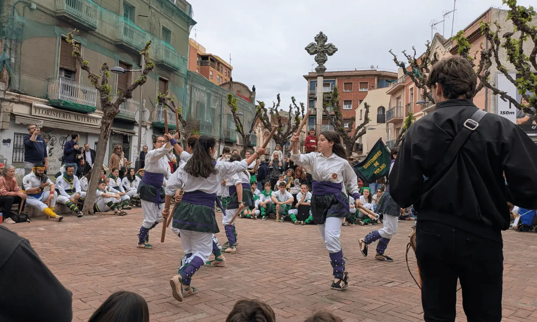 Persones participant en una dansa de bastons en una plaça urbana, amb espectadors asseguts i dempeus al voltant.