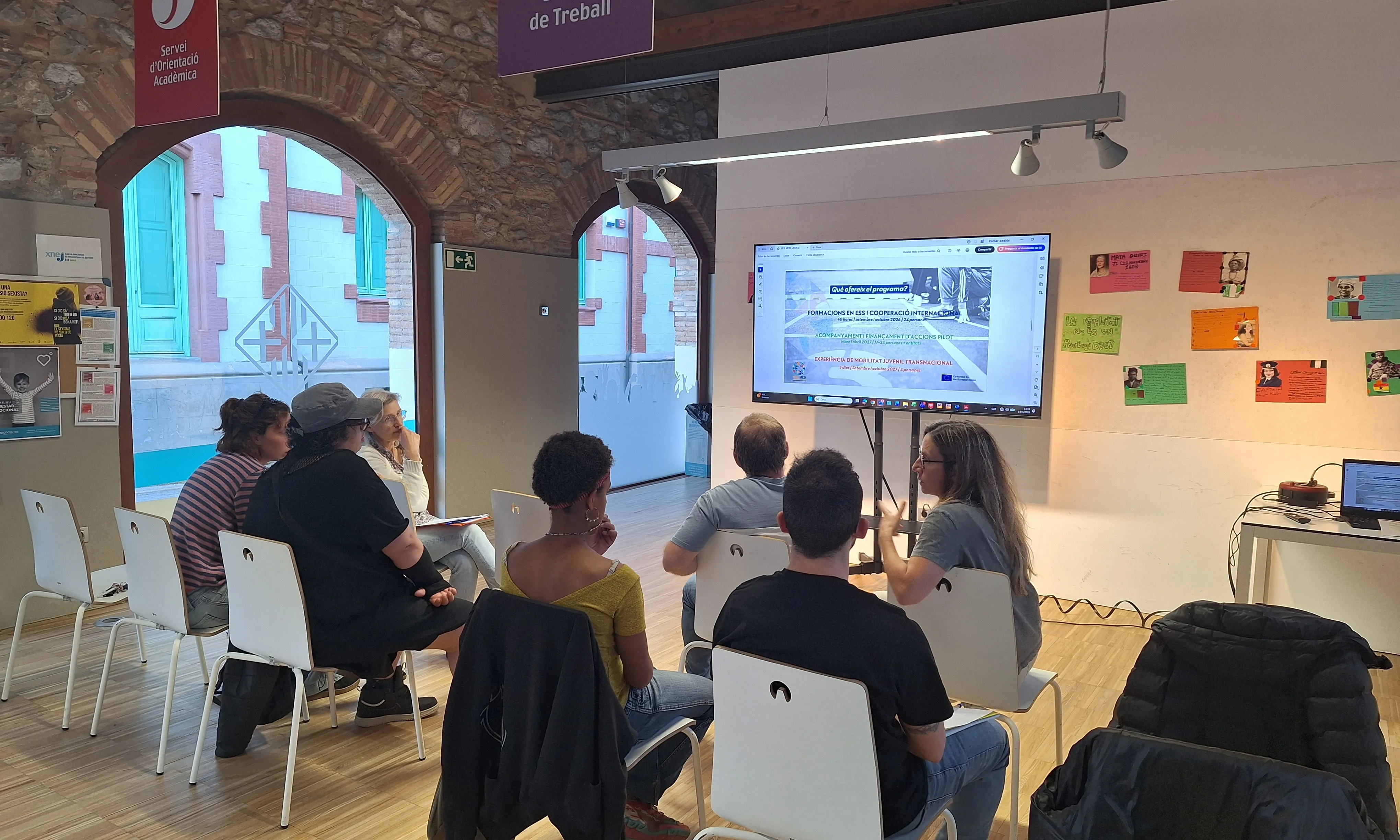 Sessió informativa del projecte. Es poden veure persones joves assegudes davant una pantalla amb informació del programa.