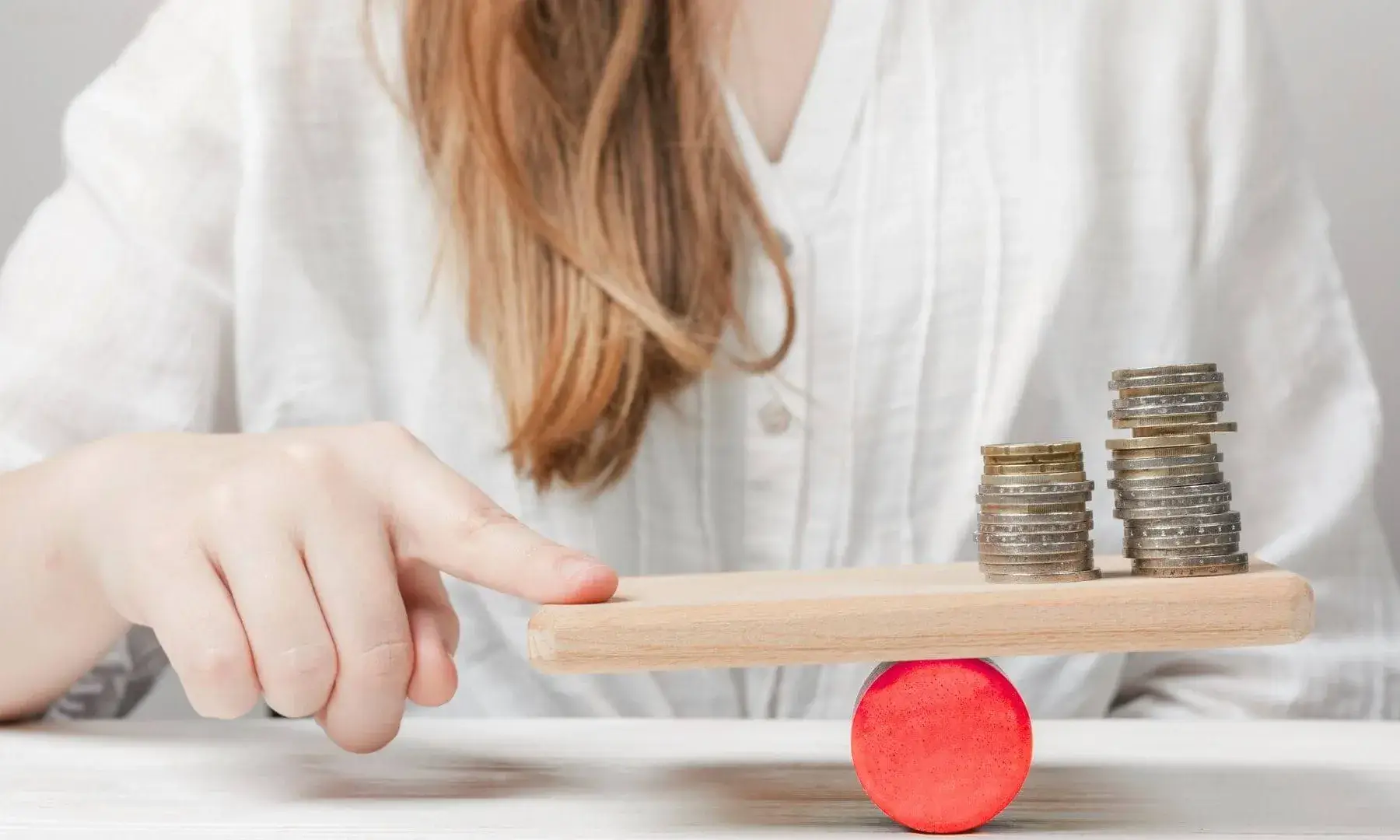 Una mà equilibra una petita fusta sobre una peça cilíndrica vermella, amb dues piles de monedes de diferents alçades damunt la fusta, simbolitzant un equilibri o comparació econòmica.