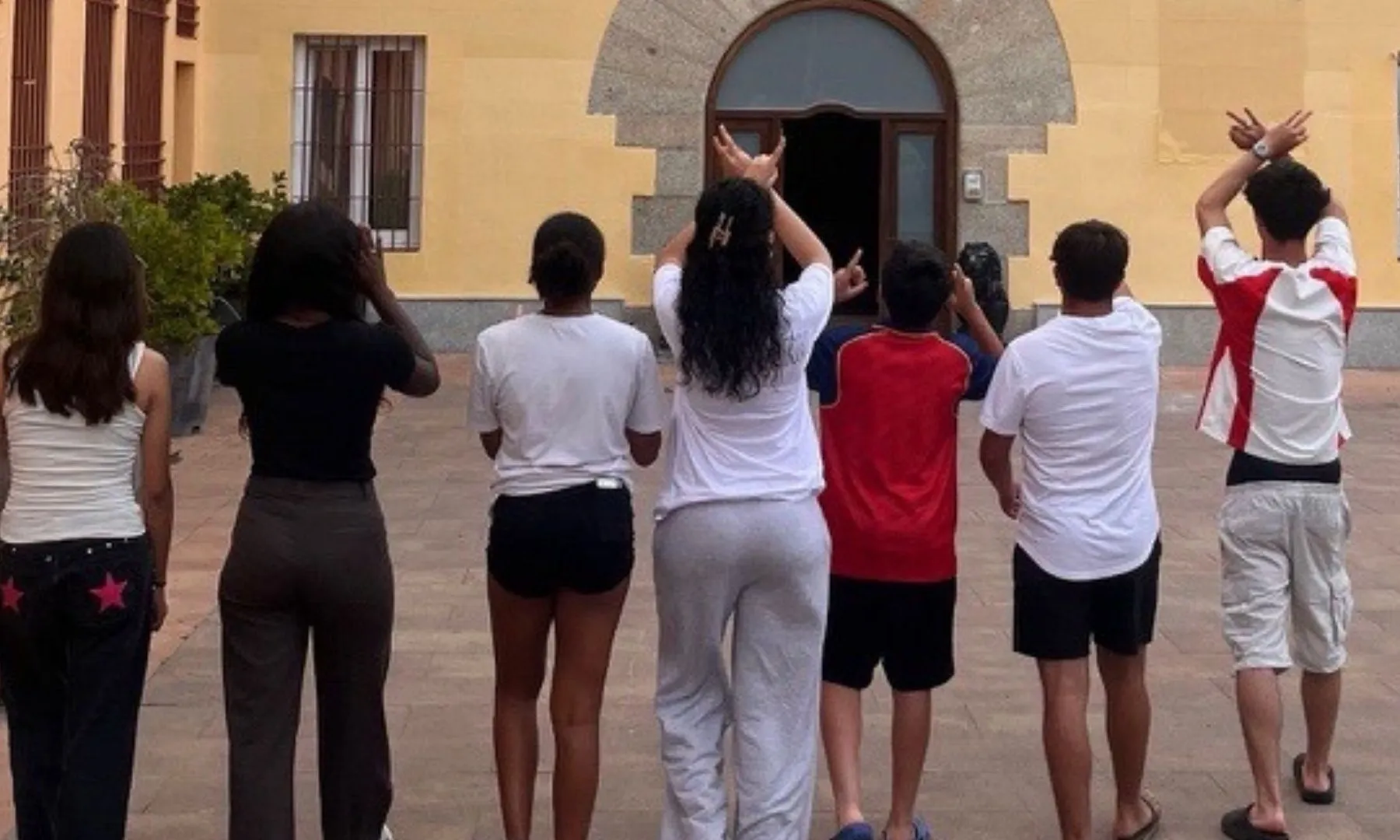 Grup d’infants i adolescents en un pati exterior, d’esquena a la càmera, participant en una activitat davant de l’entrada d’un edifici.
