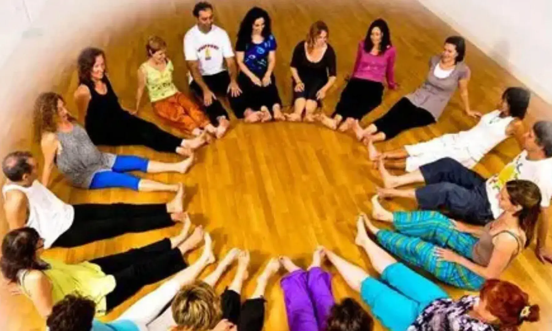 Un grup de persones adultes seuen descalces en cercle sobre un terra de fusta en una sala interior. Les participants formen una rotllana amb les cames estirades i els peus tocant-se al centre, en el marc d’una sessió grupal de moviment corporal.