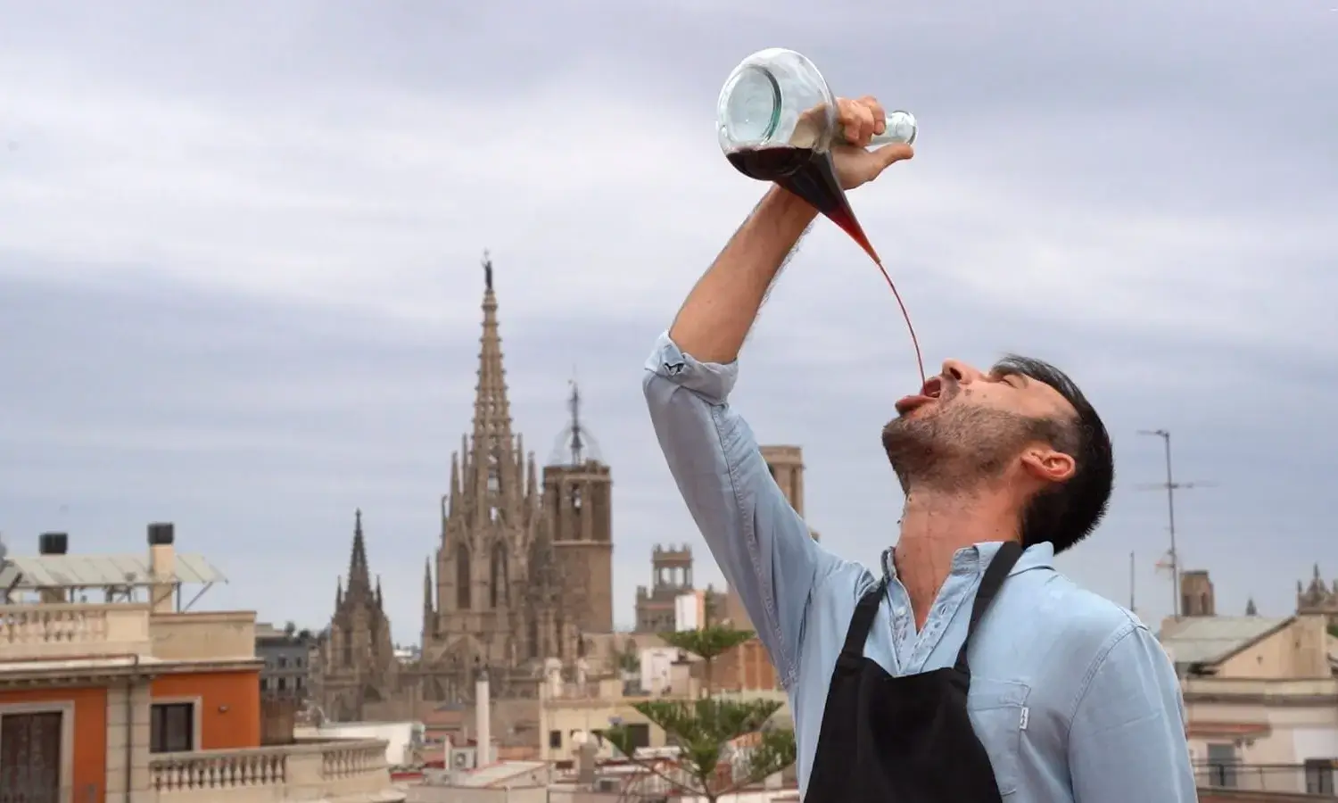 Un home veu vi d'un porró amb la Sagrada Familia de fons. 