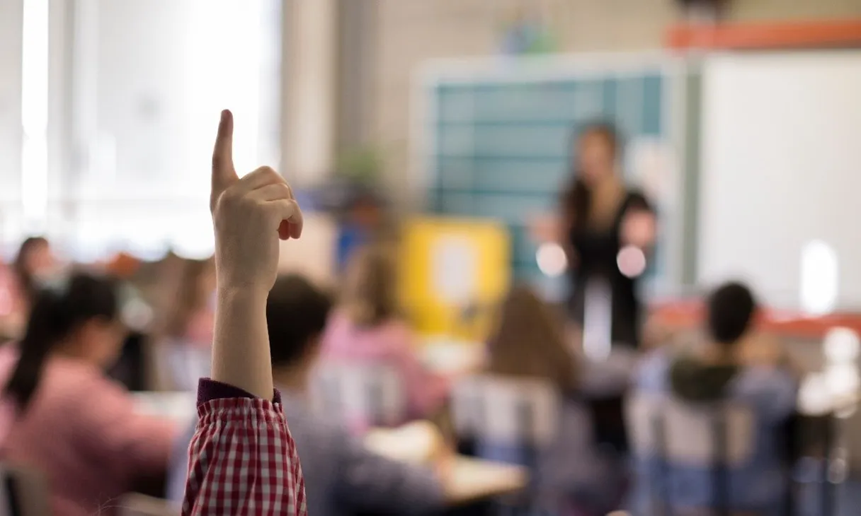 Classe on es veu un alume que alça la mà, amb la mestra de fons