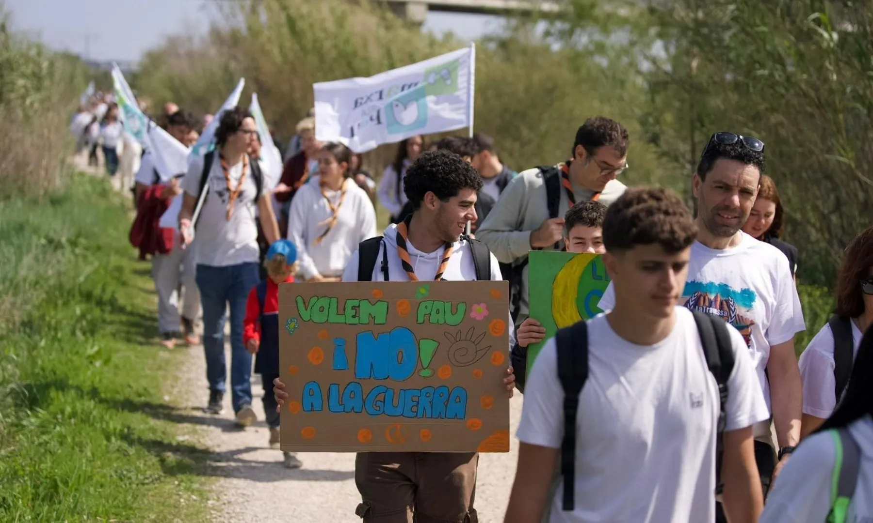 Grup de persones caminant per un camí natural amb pancartes reivindicatives per la pau durant una marxa.