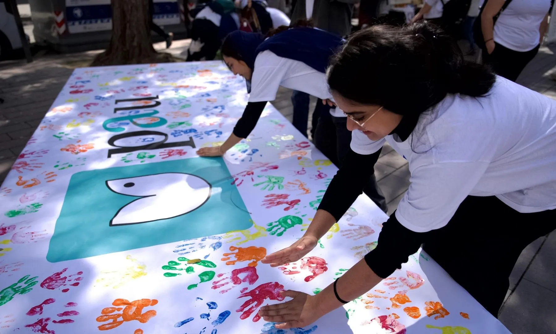 Persones posant les mans amb pintura de colors sobre un mural amb un símbol de la pau, en una activitat participativa a l’aire lliure.