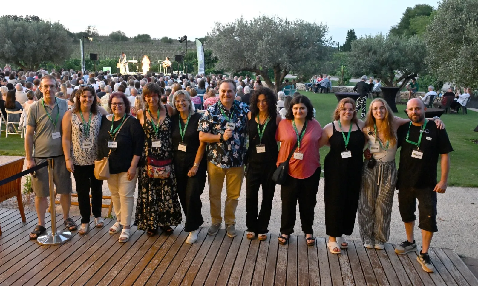 Grup de persones voluntàries. Al fons, un jardí amb oliveres on un grup de música toca davant un públic.
