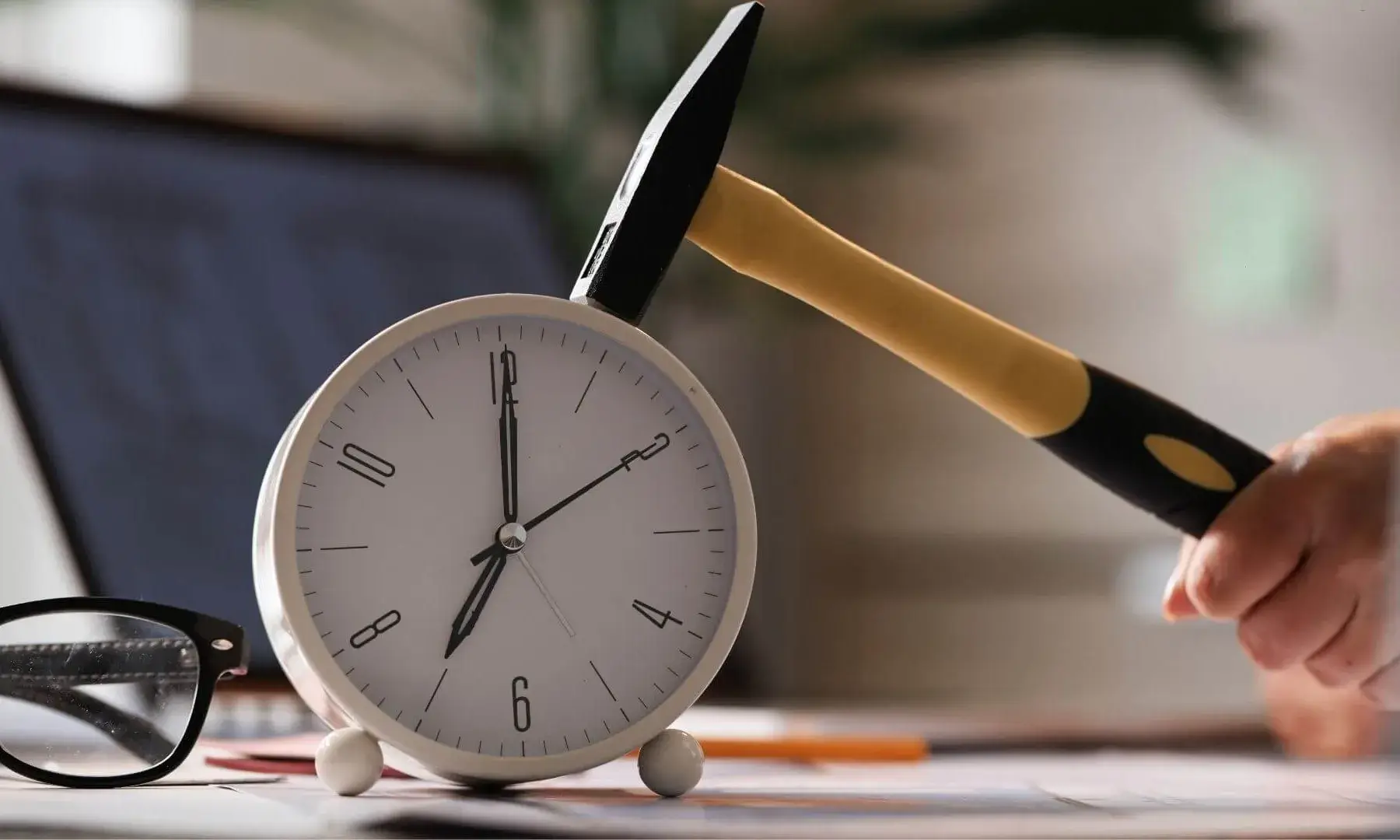 Un martell a punt de colpejar un rellotge despertador sobre una taula amb documents i ulleres.