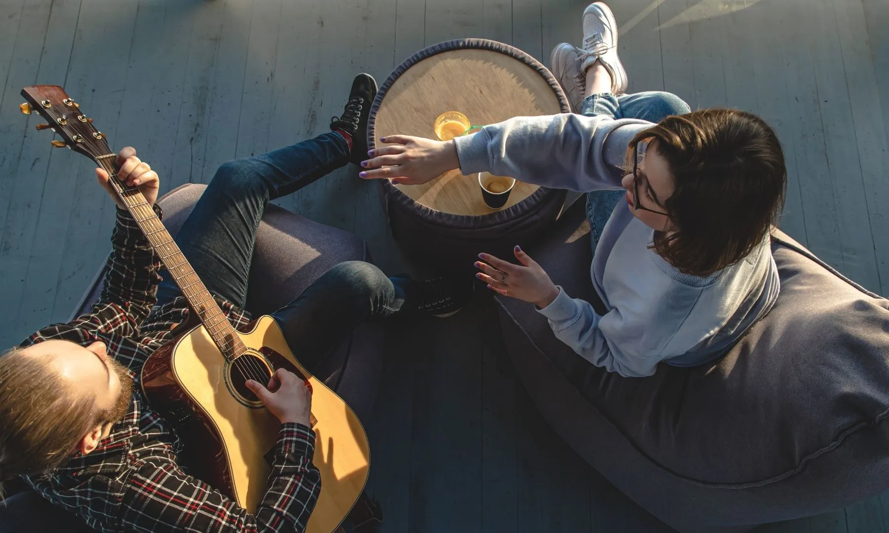 Persona tocant una guitarra mentre conversa amb una altra persona asseguda en un puf, en un ambient relaxat i informal.